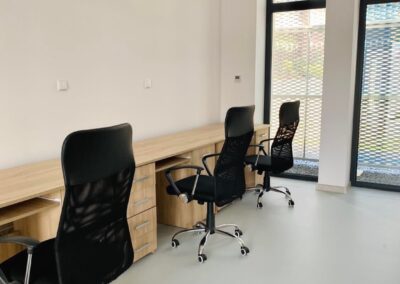 Biurka stojące przy ścianie w strefie biurowej wyposażone w czarne krzesła obrotowe. Wysokie okna przez które wpada światło dzienne. Pomieszczenie jasne, ściany w kolorze białym a podłoga w kolorze jasno szarym.