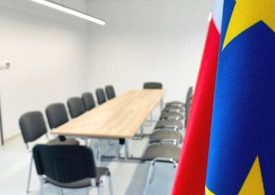 Po lewej stronie wyostrzone flagi Unii Europejskiej i Polski. W tle widać wnętrze sali konferencyjnej wyposażonej w stół w kolorze jasny dą i ustawione szare krzesła wokół. Nad stołem włączone oświetlenie.